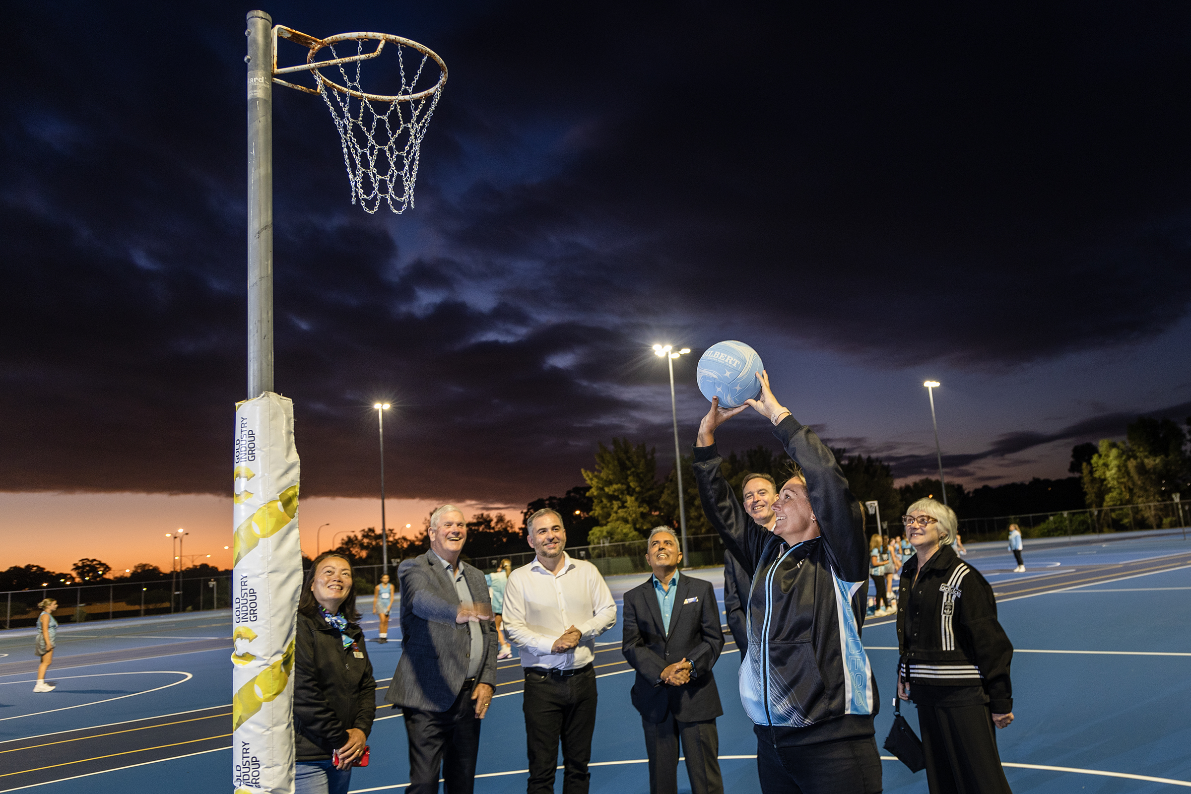 Image taken at newly updated netball courts. Six people watching as netball player shoots ball towards hoop. 
